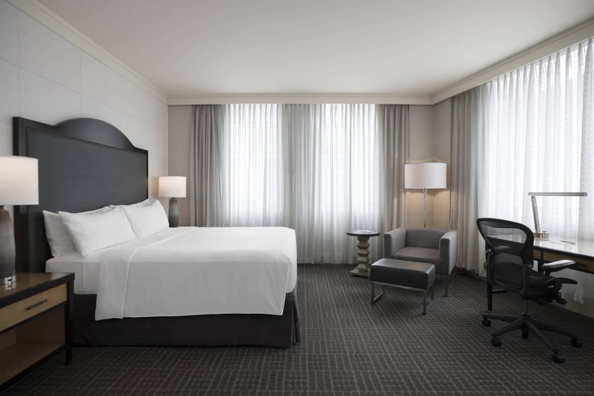 A hotel room with a large bed, two bedside tables with lamps, a desk with a chair, and a large window with curtains.