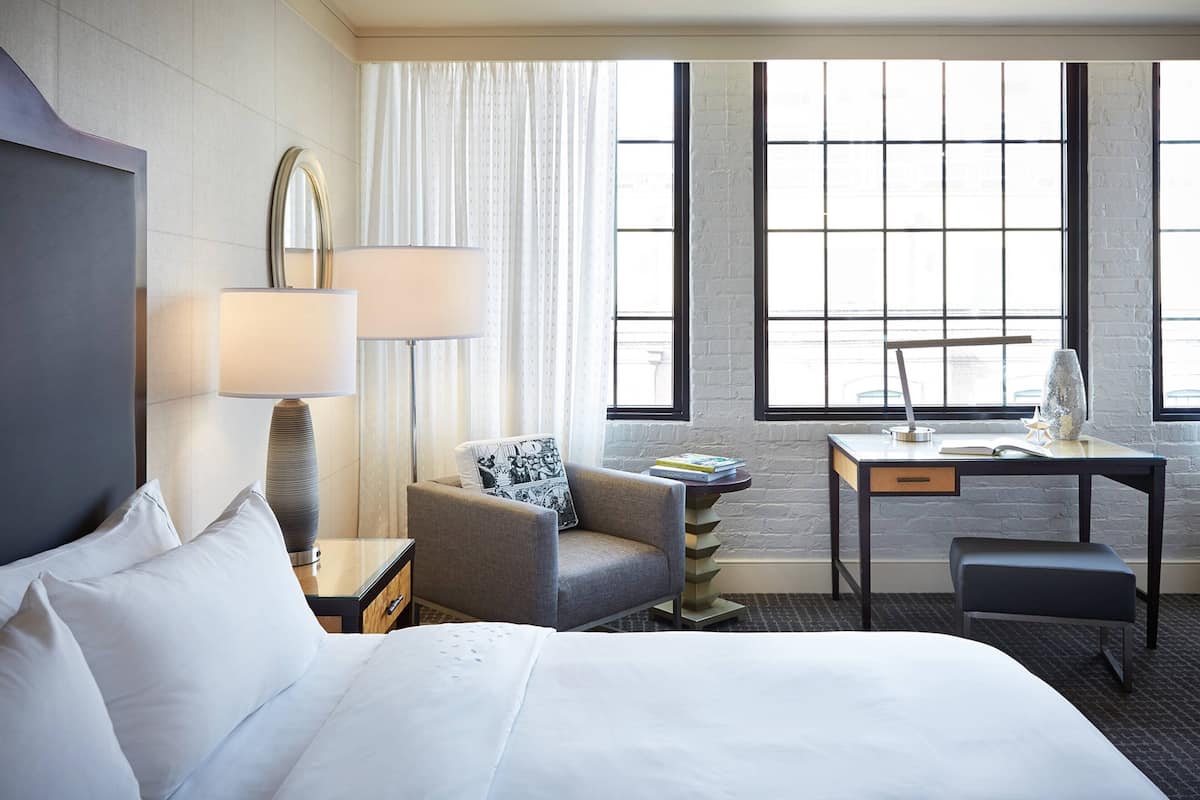 A hotel room with a bed, a desk, a chair, a lamp, and a window with curtains.