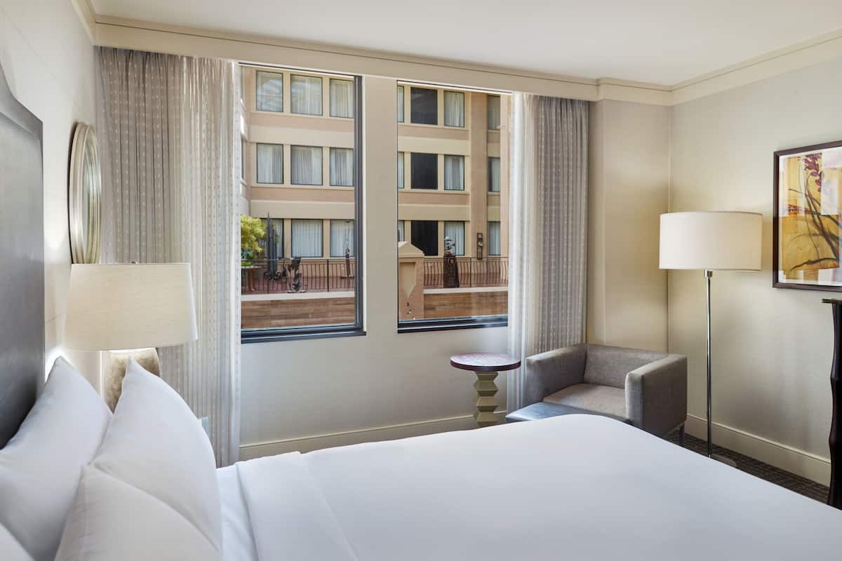 A hotel room with a large bed, a chair, a small table, and a view of the building's exterior through the windows.