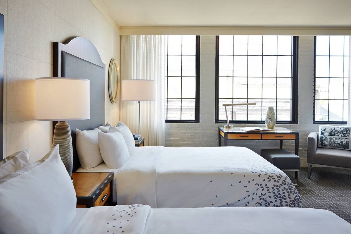 A hotel room with two beds, a desk, a chair, and large windows.