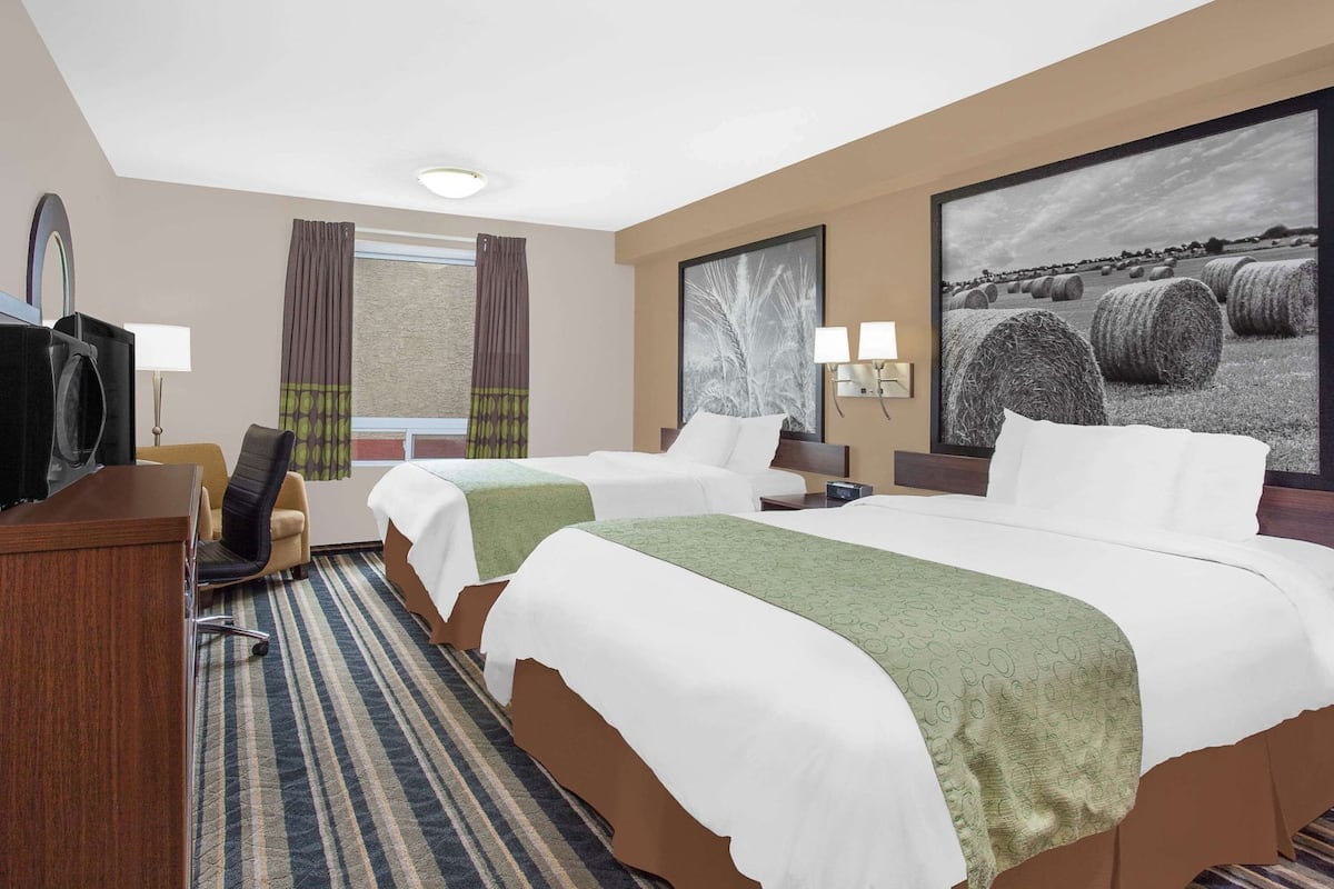 A hotel room with two beds, a desk with a television, a chair, a window with curtains, and a large painting on the wall.