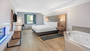 A modern hotel room with a large bed, a desk with a flat-screen TV, a desk lamp, a window with curtains, a wall-mounted TV, a built-in bathtub, and a small heater. - Super 8 by Wyndham Cornwall ON (Cornwall)