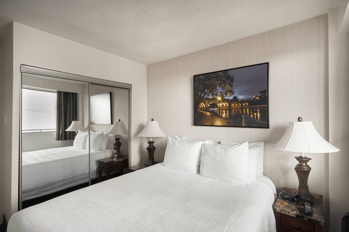 Quarto de hotel com uma cama grande, um espelho, uma mesa de cabeceira com abajur e um quadro na parede.