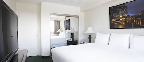 A hotel room with a large bed, a flat-screen TV, a mirror, a painting, and a nightstand with a lamp.