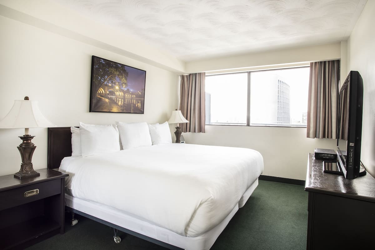 Quarto de hotel com uma cama grande, duas mesas de cabeceira com luminárias, uma televisão de tela plana e uma janela com cortinas.