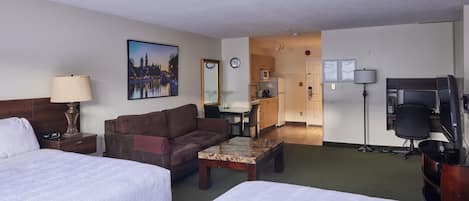 Habitación de hotel con dos camas, un sofá, una mesa de centro, un escritorio con una computadora, un televisor y una cocina al fondo.
