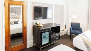 Habitación de hotel con cama, televisor, minibar, una silla y una mesita de noche con lámpara.