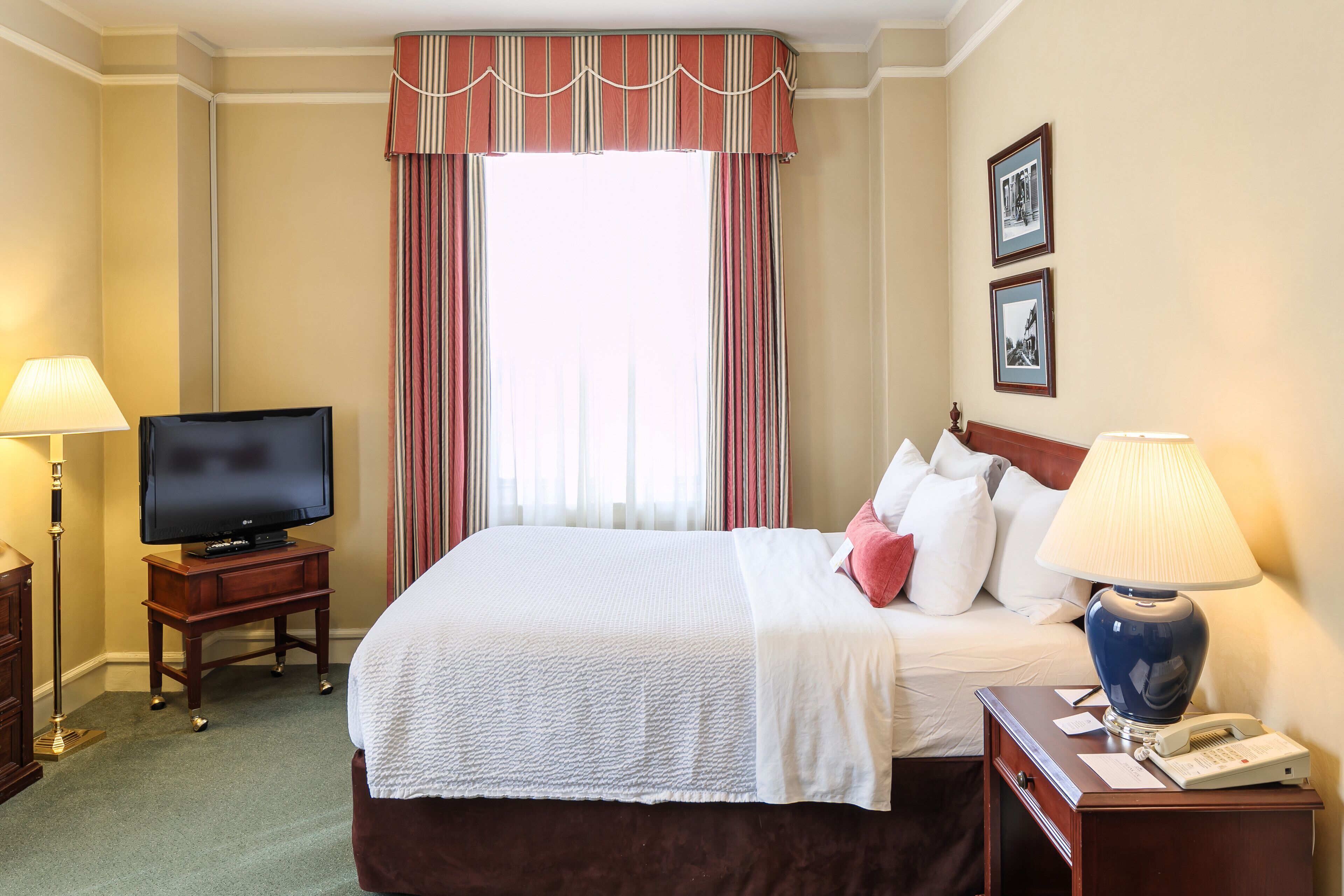Una habitación de hotel con cama, televisión, lámpara, teléfono y ventana con cortinas.