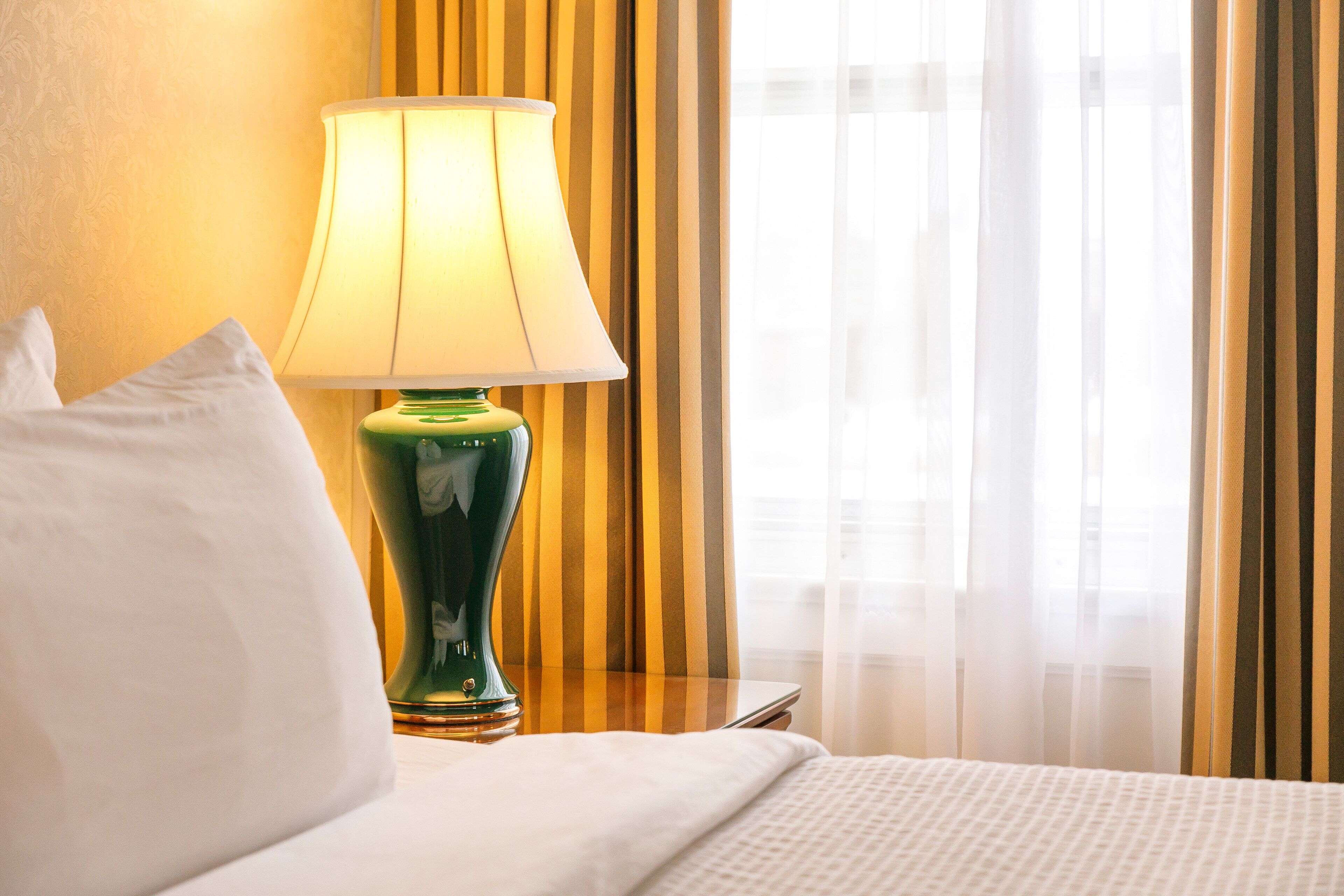 A neatly made bed with white linens, a green table lamp, and a striped curtain in the background.