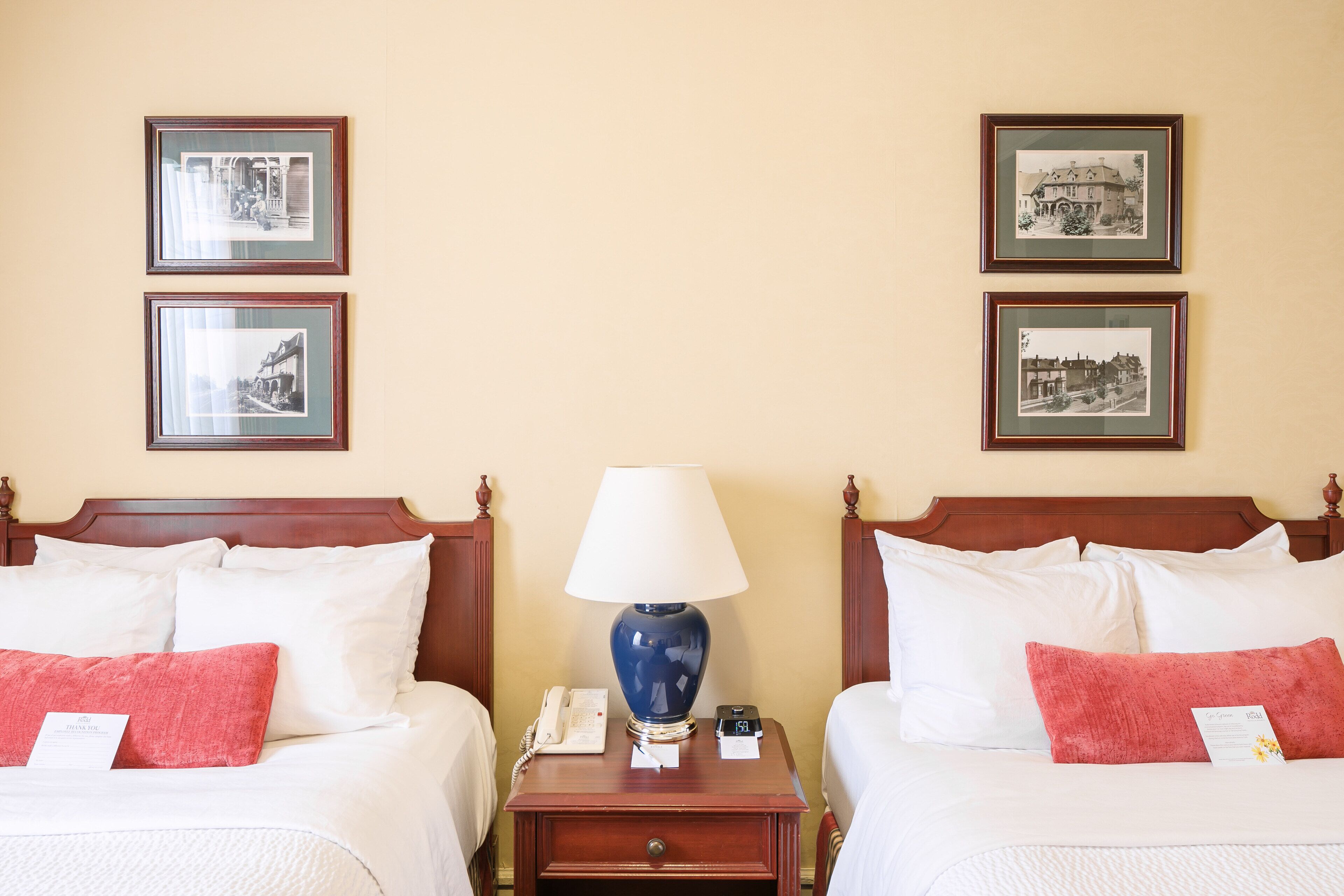 A hotel room with two beds, a nightstand with a lamp, a phone, and a digital clock. There are framed pictures on the wall.