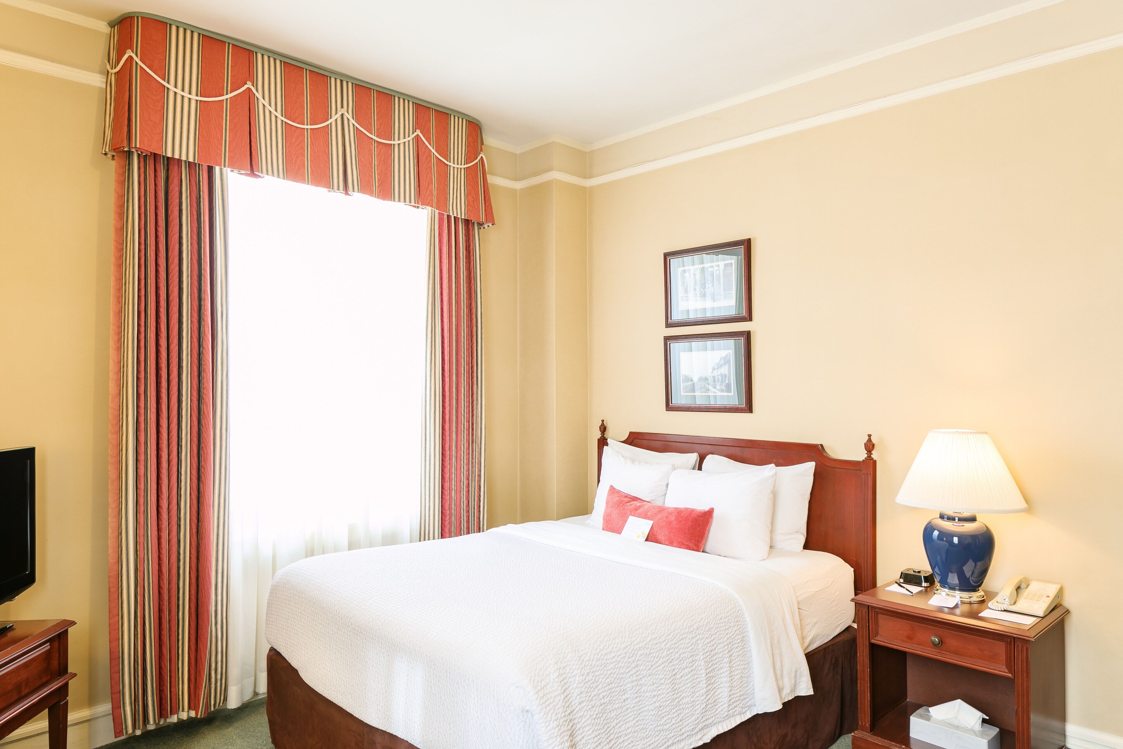 A hotel room with a large bed, a nightstand with a lamp, a television, and a window with curtains.