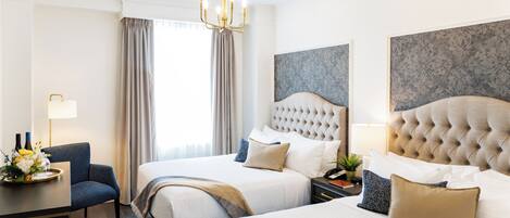 A hotel room with two beds, a chandelier, a desk with a lamp, and a large window with curtains.