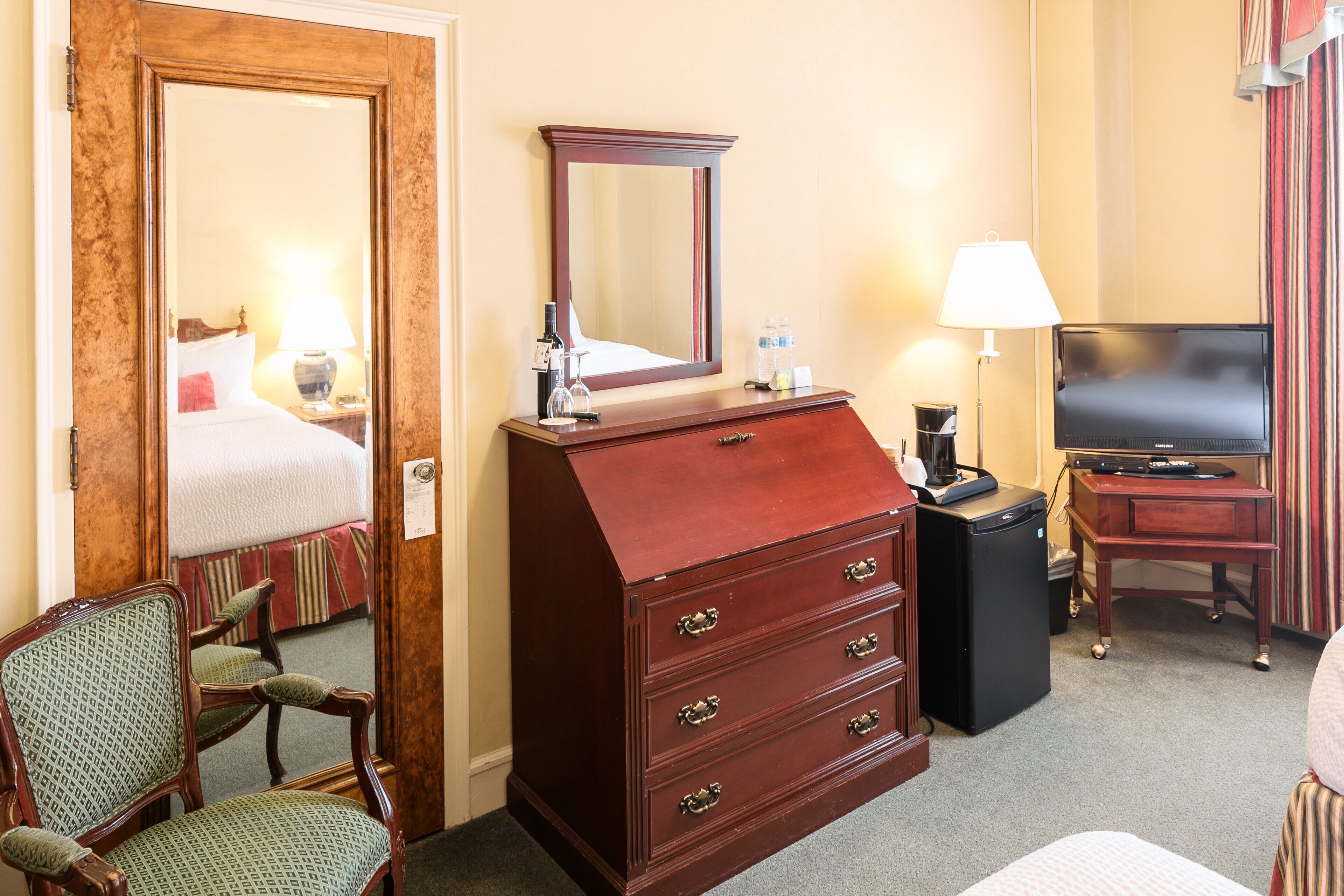 A hotel room with a wooden dresser, a chair, a television, and a desk with a lamp.