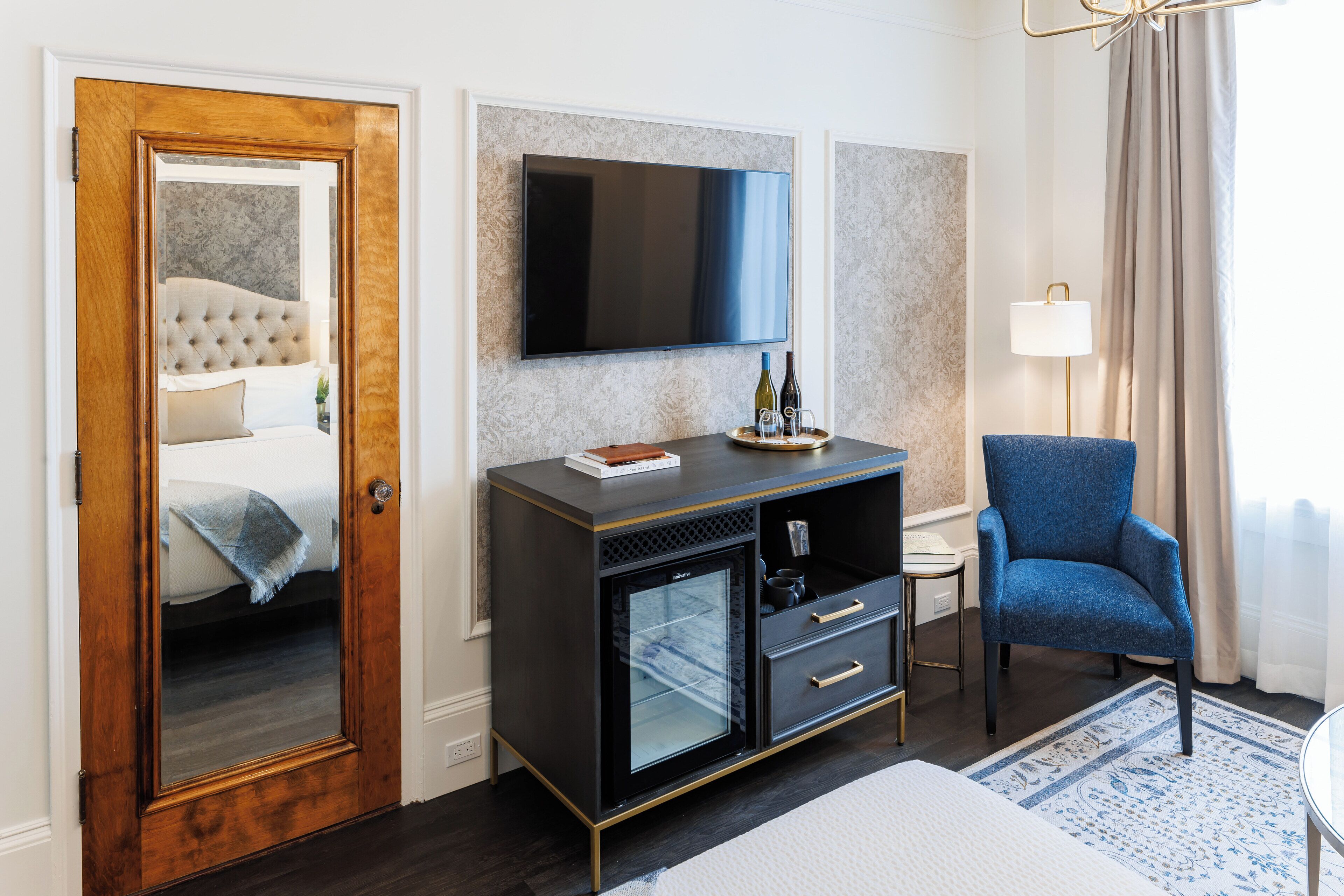 A hotel room with a wooden door, a bed with a tufted headboard, a flat-screen TV, a minibar, a blue armchair, and a patterned rug.
