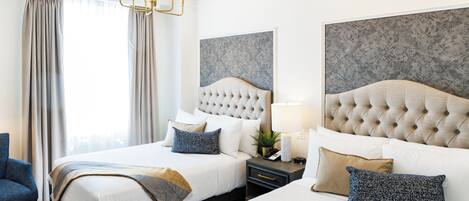 A hotel room with two beds, a chandelier, a large window with curtains, and a headboard with a floral pattern.