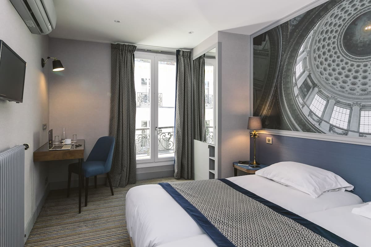 A hotel room with a bed, a desk with a laptop, a chair, a window with curtains, a lamp, and a large wall mural.