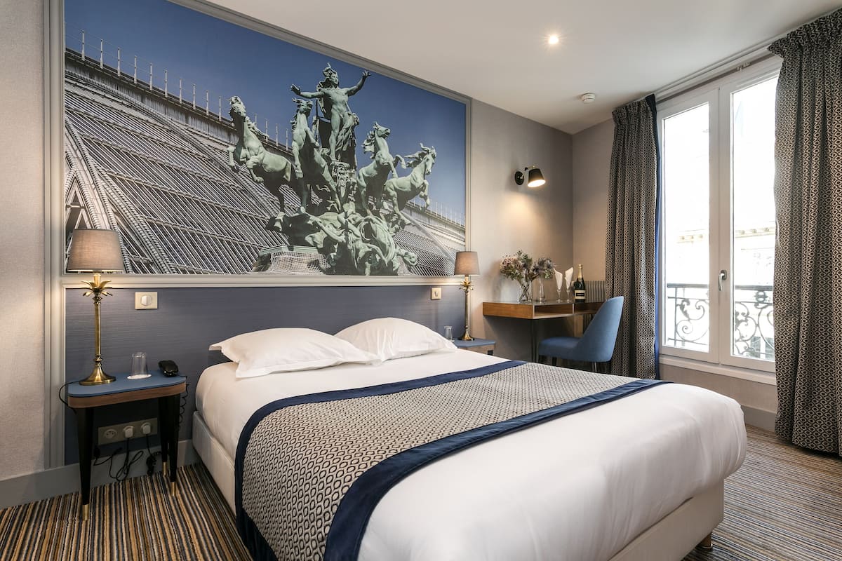 A hotel room with a large bed, a nightstand with a lamp, a desk with a chair, a window with curtains, and a painting of a historical scene on the wall.