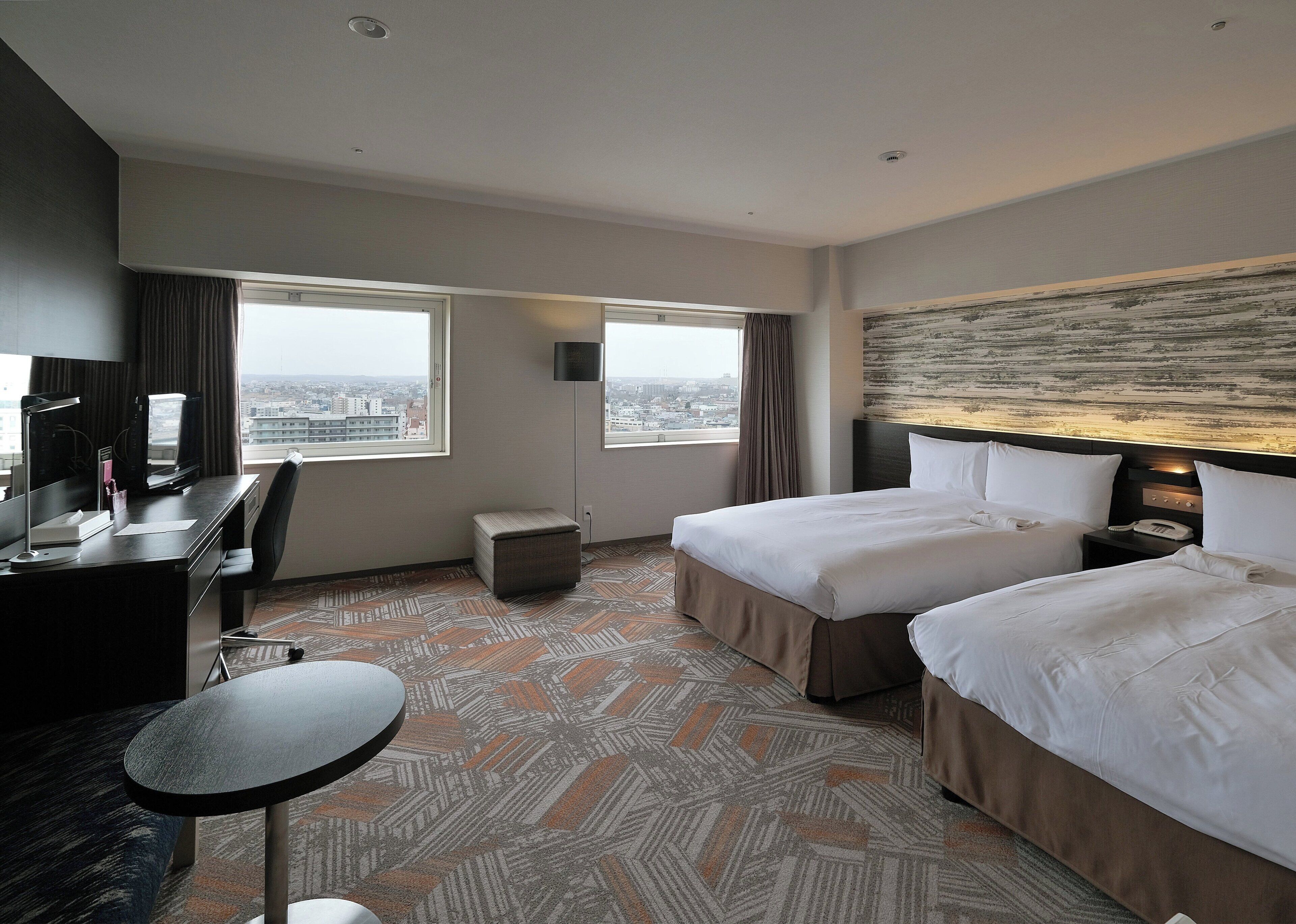 A hotel room with a desk, two beds, a small table, and a city view.