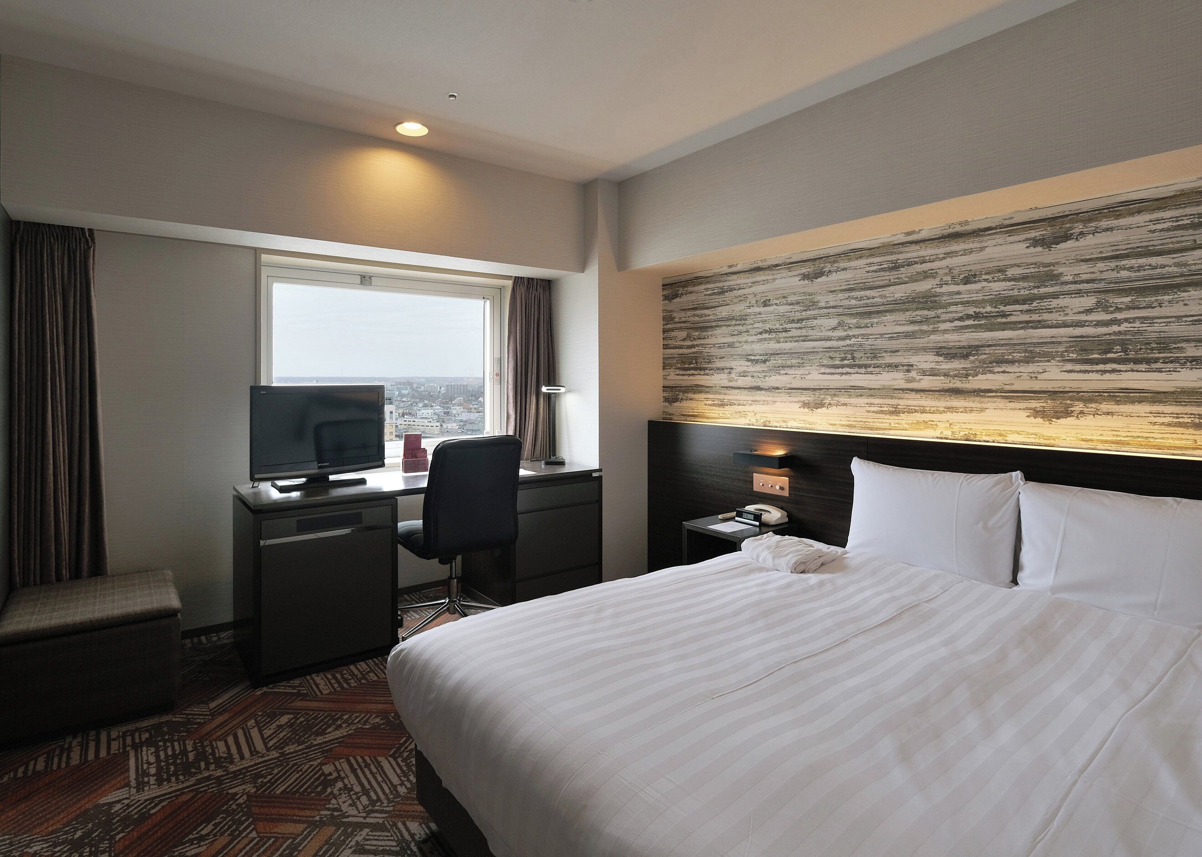 Ein Hotelzimmer mit Bett, Schreibtisch, Stuhl, Fernseher und Blick auf die Stadt.