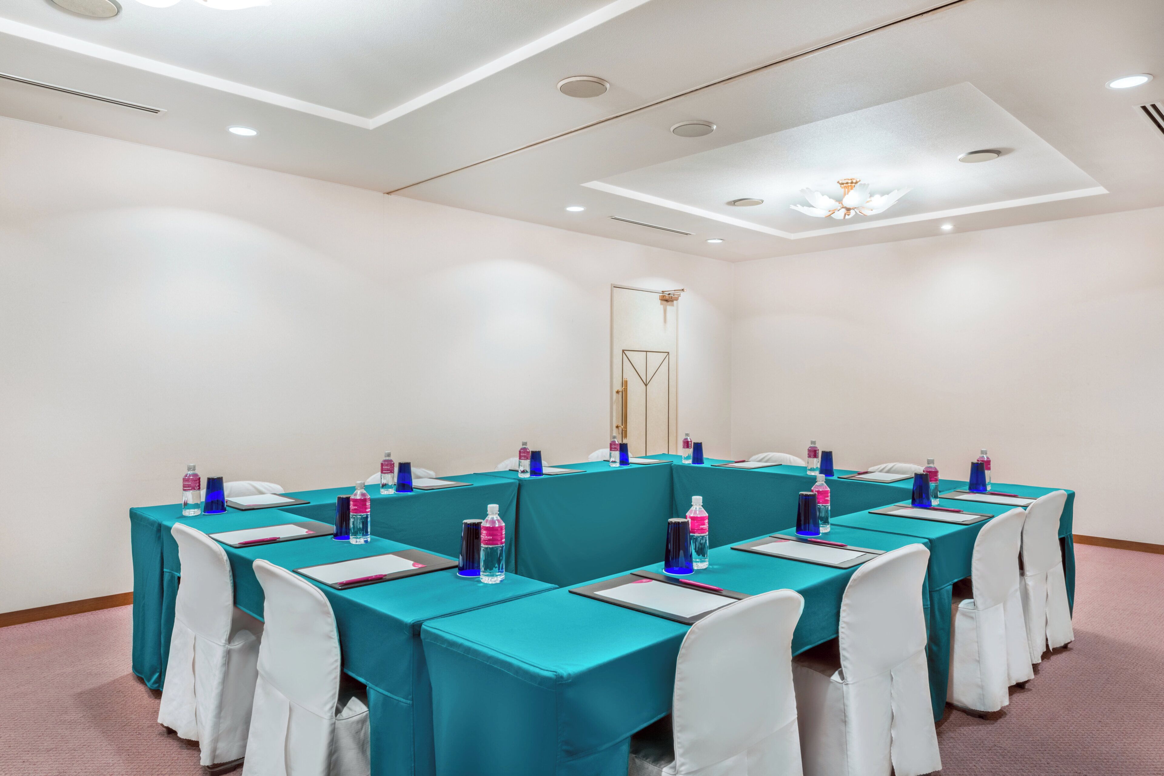 A conference room with teal tables and white chairs, equipped with water bottles and notepads.