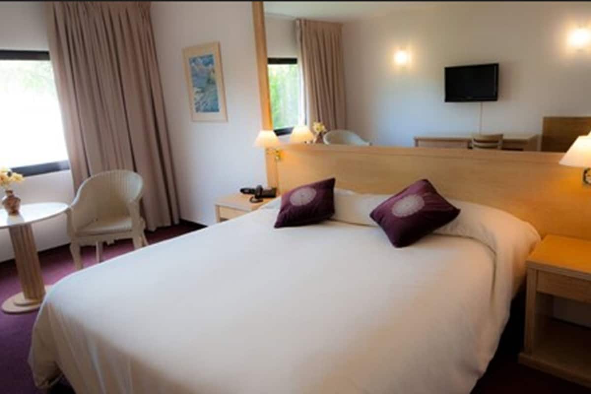Una habitación de hotel con una cama grande, dos mesitas de noche, una silla, una mesita pequeña, un televisor y una ventana con cortinas.