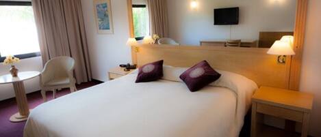 A hotel room with a large bed, two bedside tables, a chair, a small table, a TV, and a window with curtains.