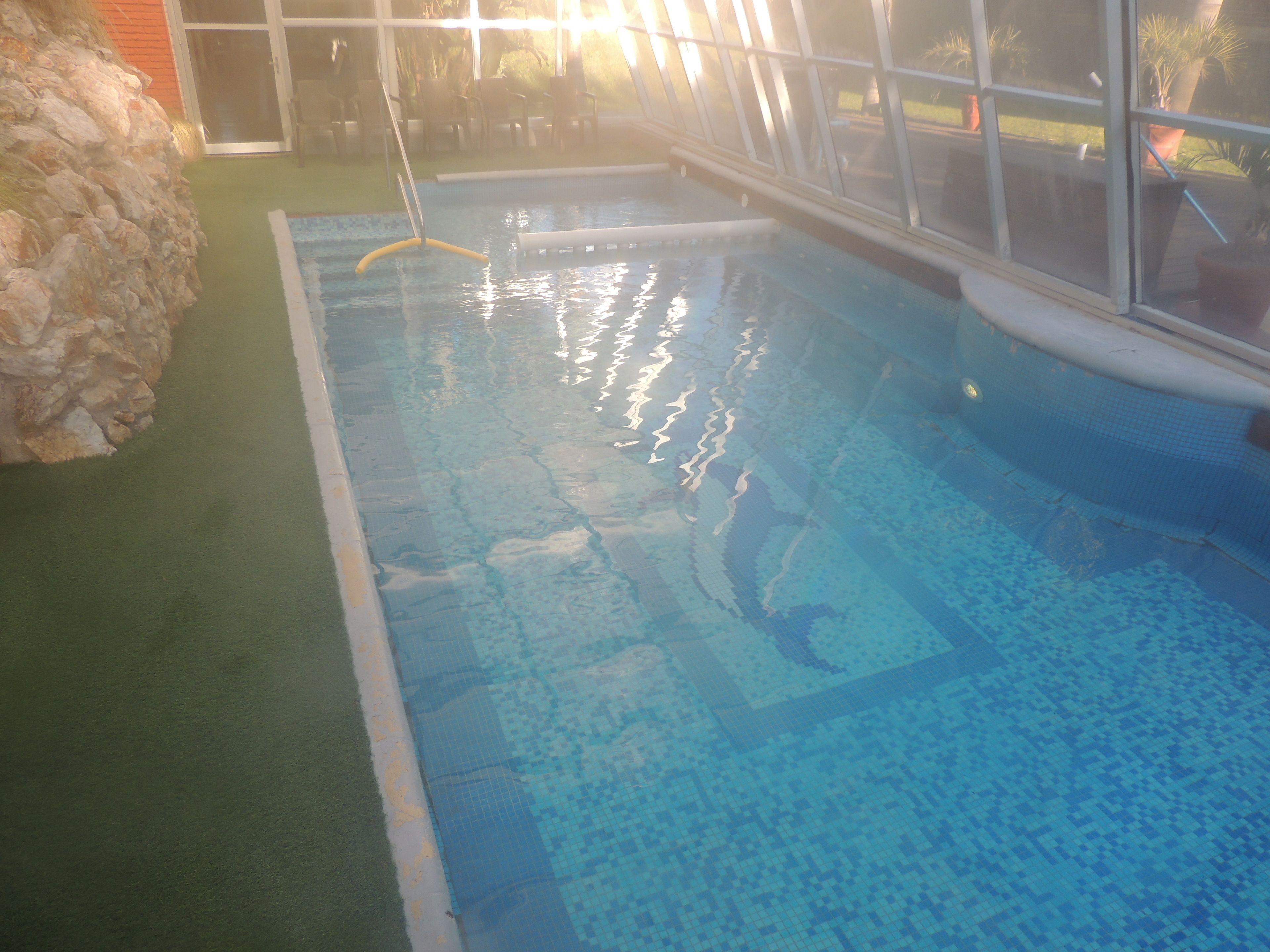 An indoor swimming pool with clear blue water, a stone wall on one side, and a glass door leading to an outdoor area.