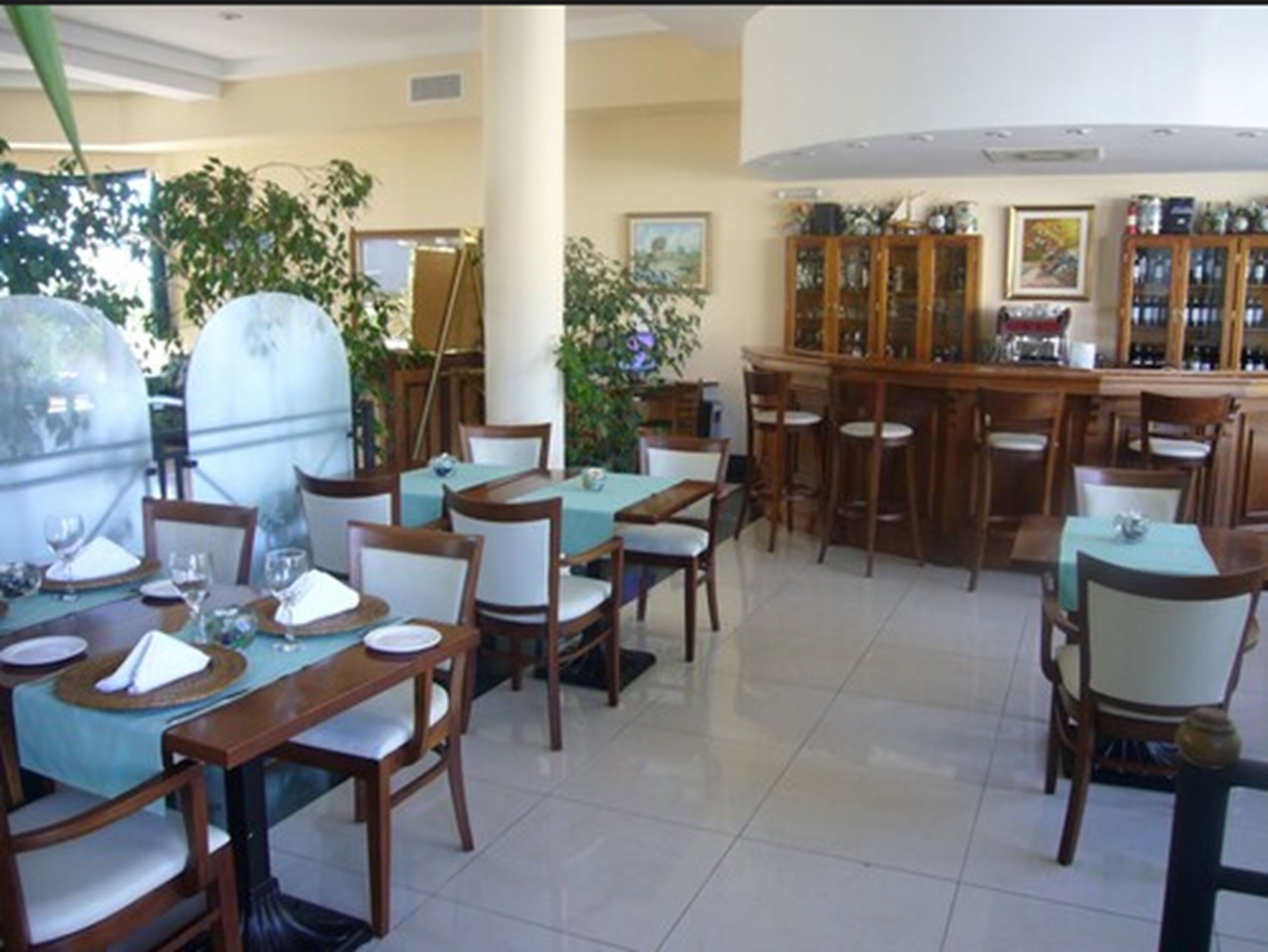 A dining area with wooden tables and chairs, a bar area with stools, and a large window with plants.
