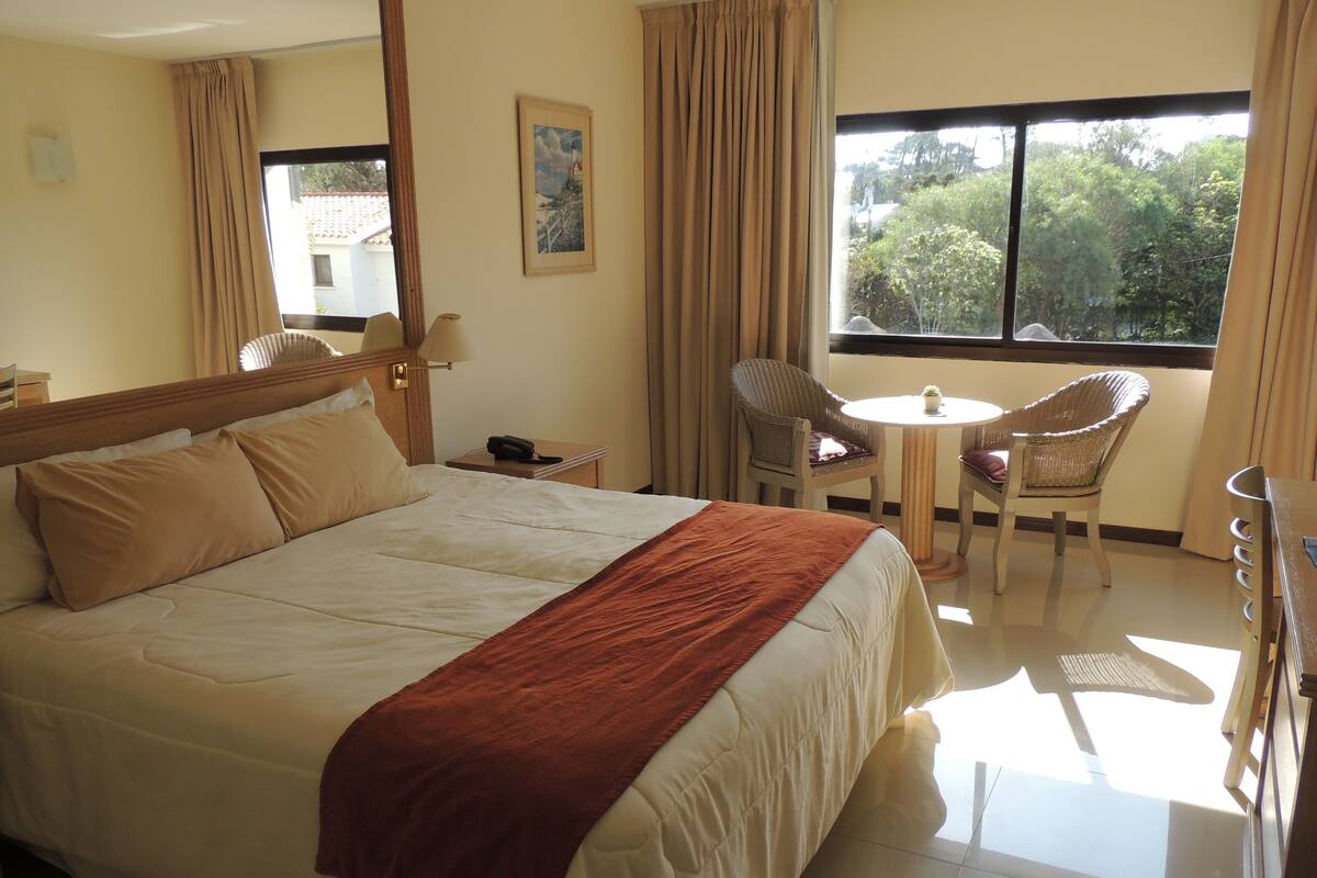 Habitación de hotel con una cama grande, una mesita con sillas, una mesita de noche y una ventana con cortinas.