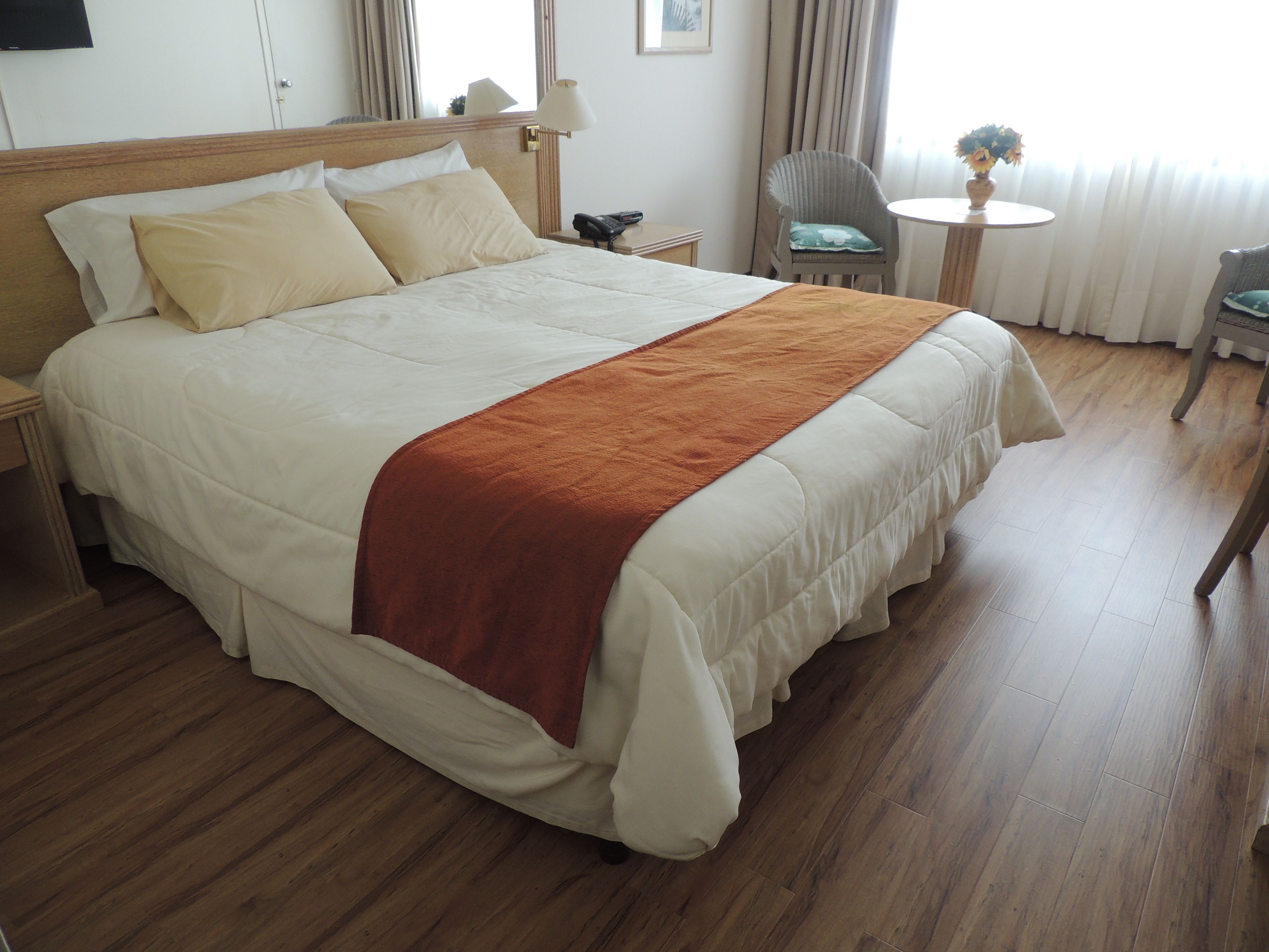 Habitación de hotel con una cama grande, una mesita de noche, una silla, una mesa redonda pequeña y un ventanal con cortinas.