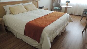 Habitación de hotel con una cama grande, una mesita de noche, una silla, una mesa redonda pequeña y un ventanal con cortinas.