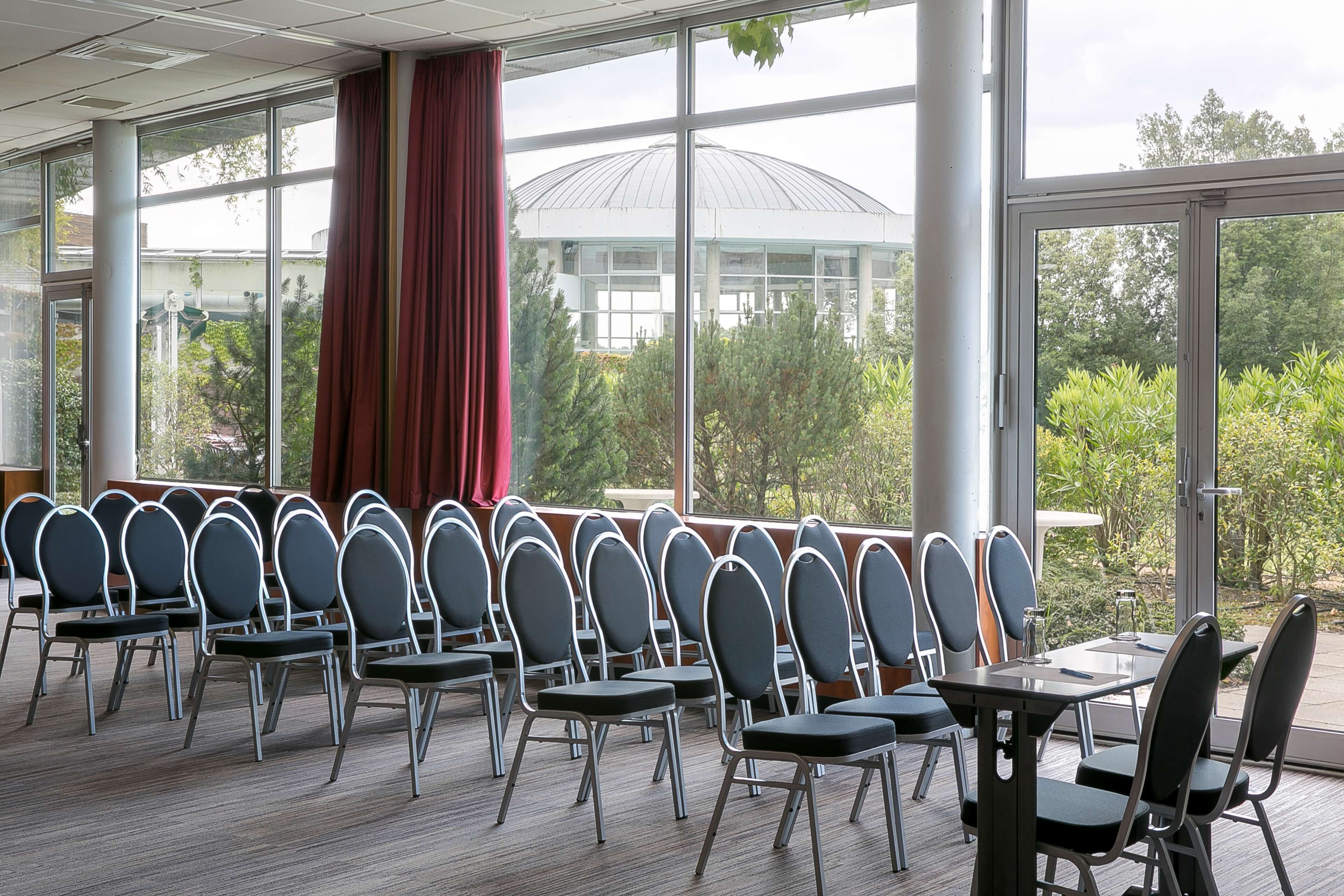 Un salón de conferencias con filas de sillas y mesas, ventanas amplias y vistas a zonas verdes y un edificio al fondo.