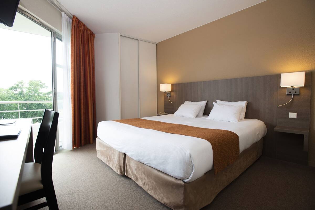 A hotel room with a large bed, two bedside lamps, a desk, and a view of greenery outside.