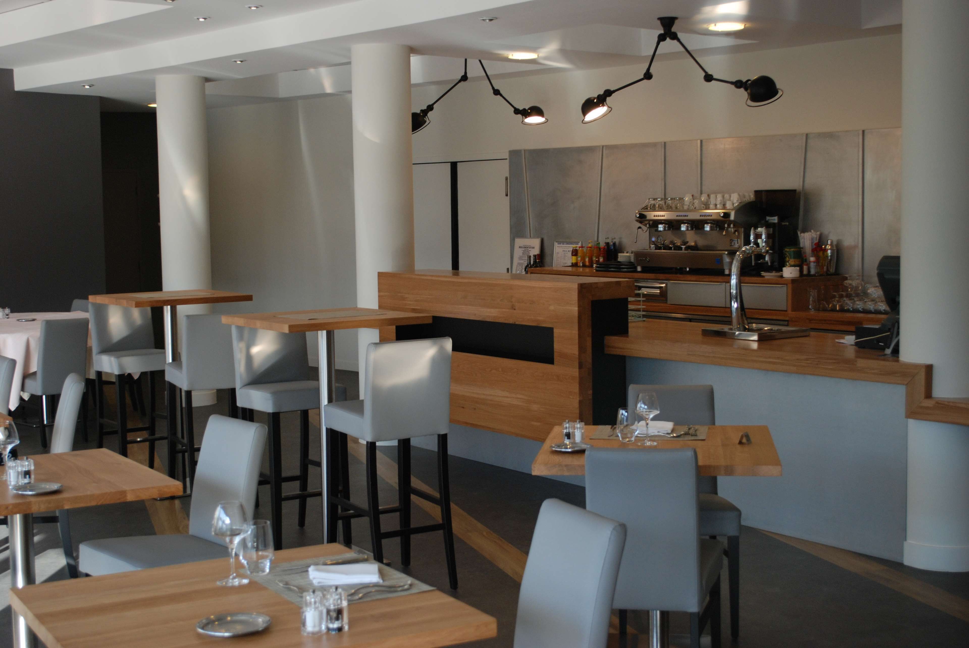 Ein modernes Restaurant mit Holztischen und grauen Stühlen, einem Barbereich mit Theke und Pendelleuchten.