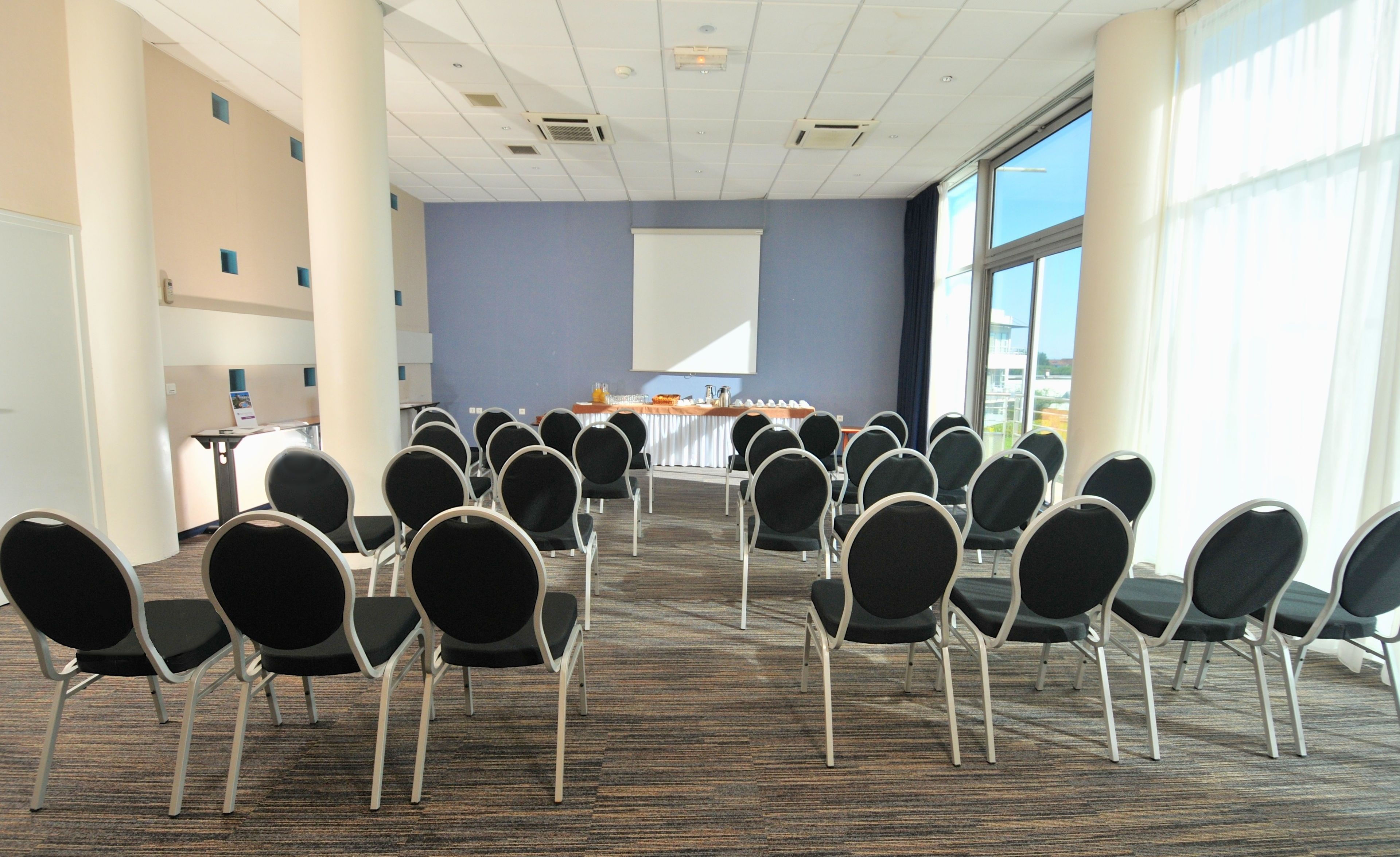 Un salón de conferencias con filas de sillas orientadas hacia una pantalla de presentación, amplias ventanas y suelo alfombrado.