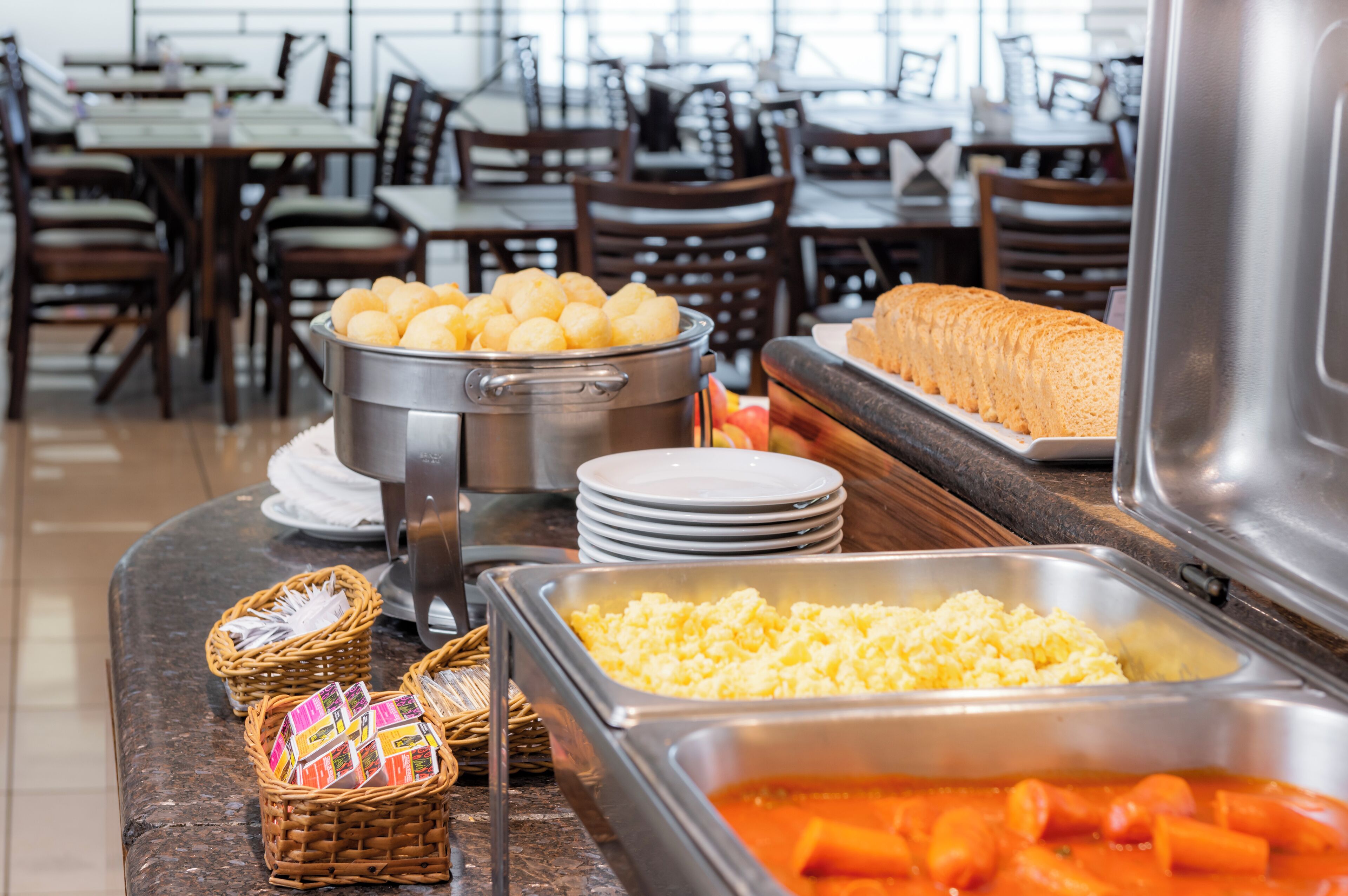 A buffet setup with various food items, including a pot of potatoes, a tray of bread, and a container of scrambled eggs.