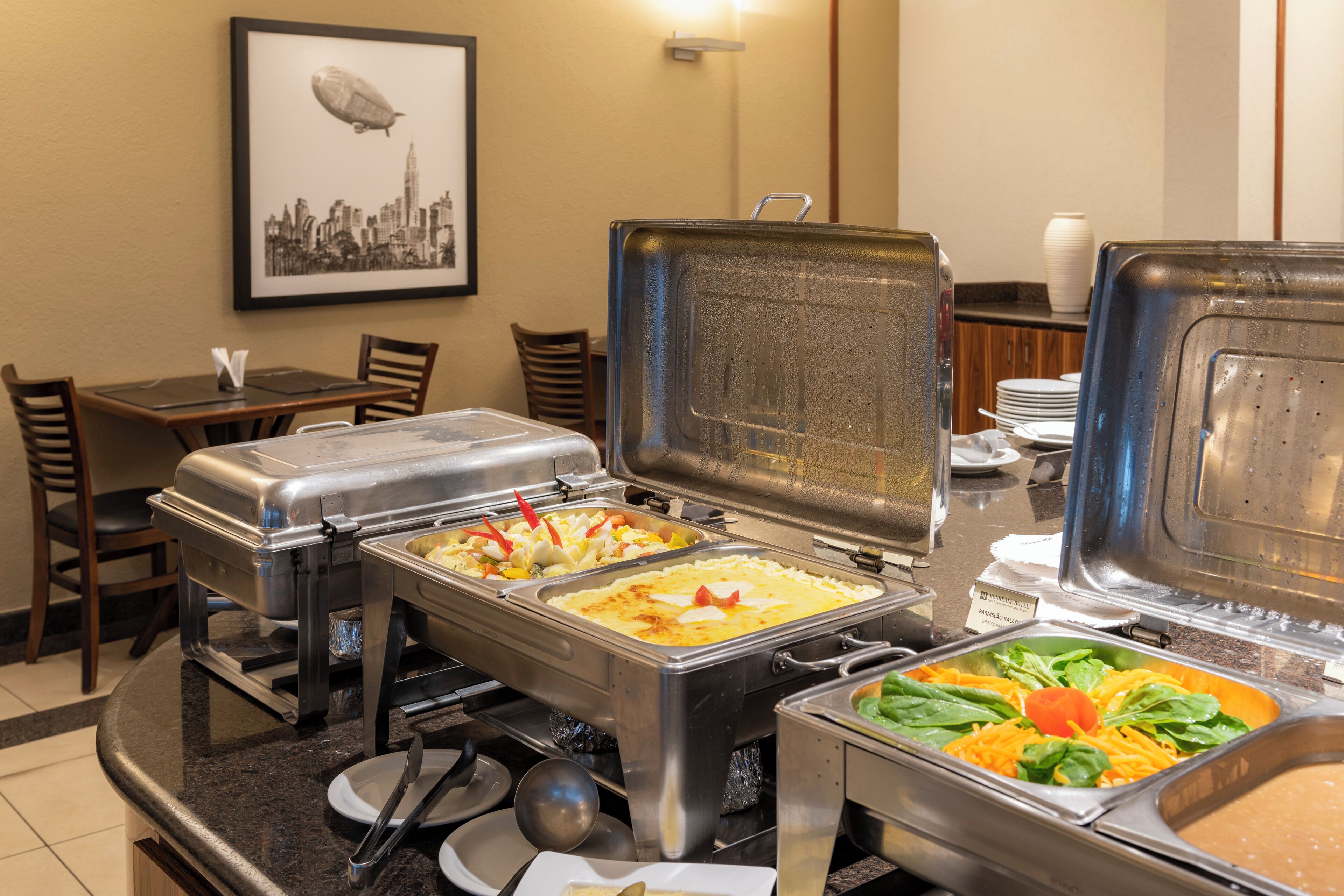 A buffet setup with various food items in metal containers, a framed cityscape picture on the wall, and a table with chairs.