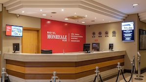 Ein Hotelrezeptionstresen mit roter Wand, einem digitalen Bildschirm, der Hotelinformationen anzeigt, und einer Uhr.