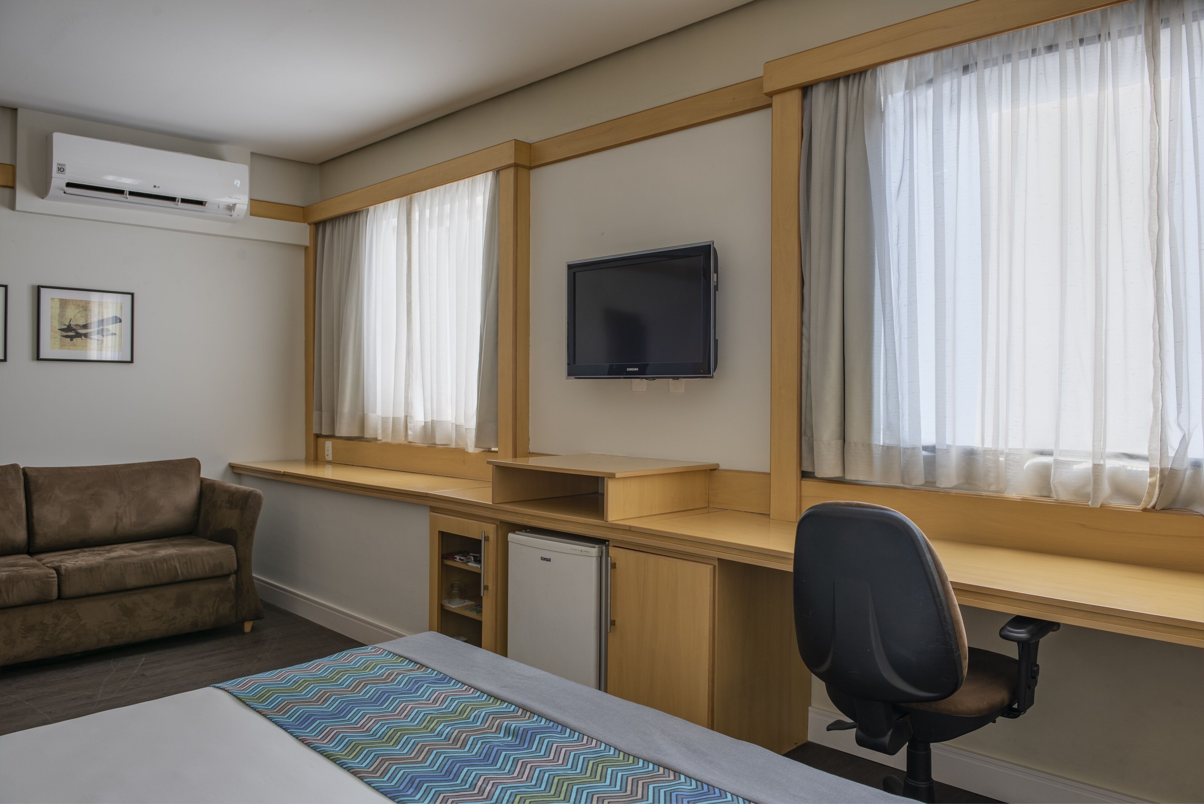 Habitación de hotel con cama, escritorio, silla, televisor, sofá y un refrigerador pequeño.