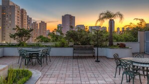 Eine Dachterrasse mit Sitzgelegenheiten im Freien, umgeben von Pflanzen und mit Blick auf eine Stadtlandschaft bei Sonnenuntergang.