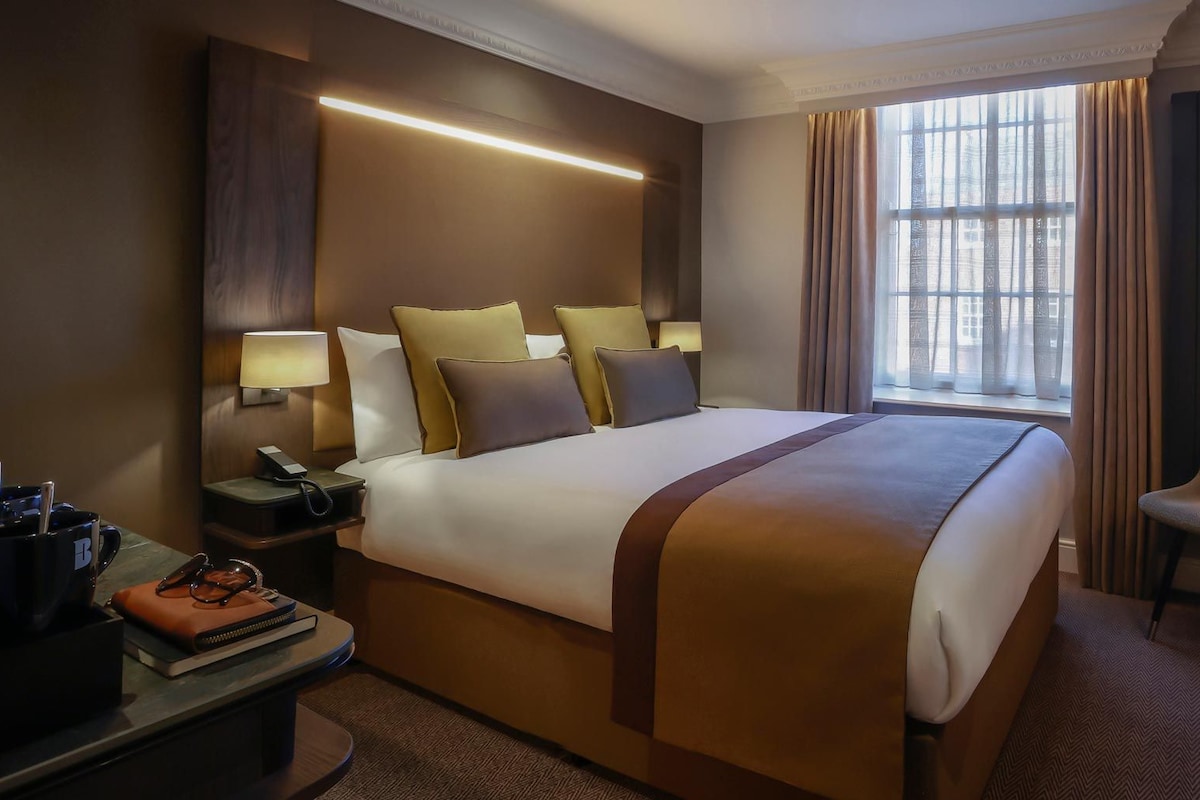 A hotel room with a large bed, bedside lamps, a desk with a phone and a mug, a chair with a handbag, and a TV mounted on the wall.
