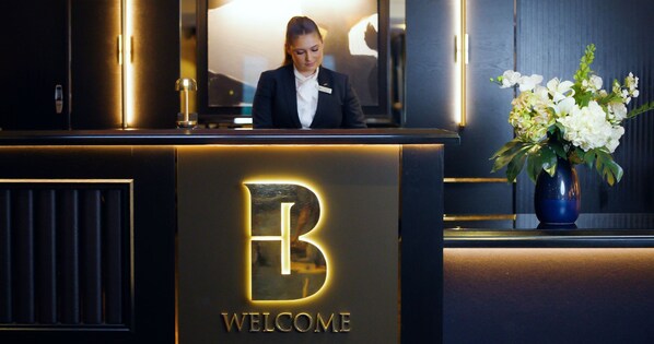 A reception desk with a 'B' logo and 'WELCOME' text, a receptionist, and a vase of flowers.