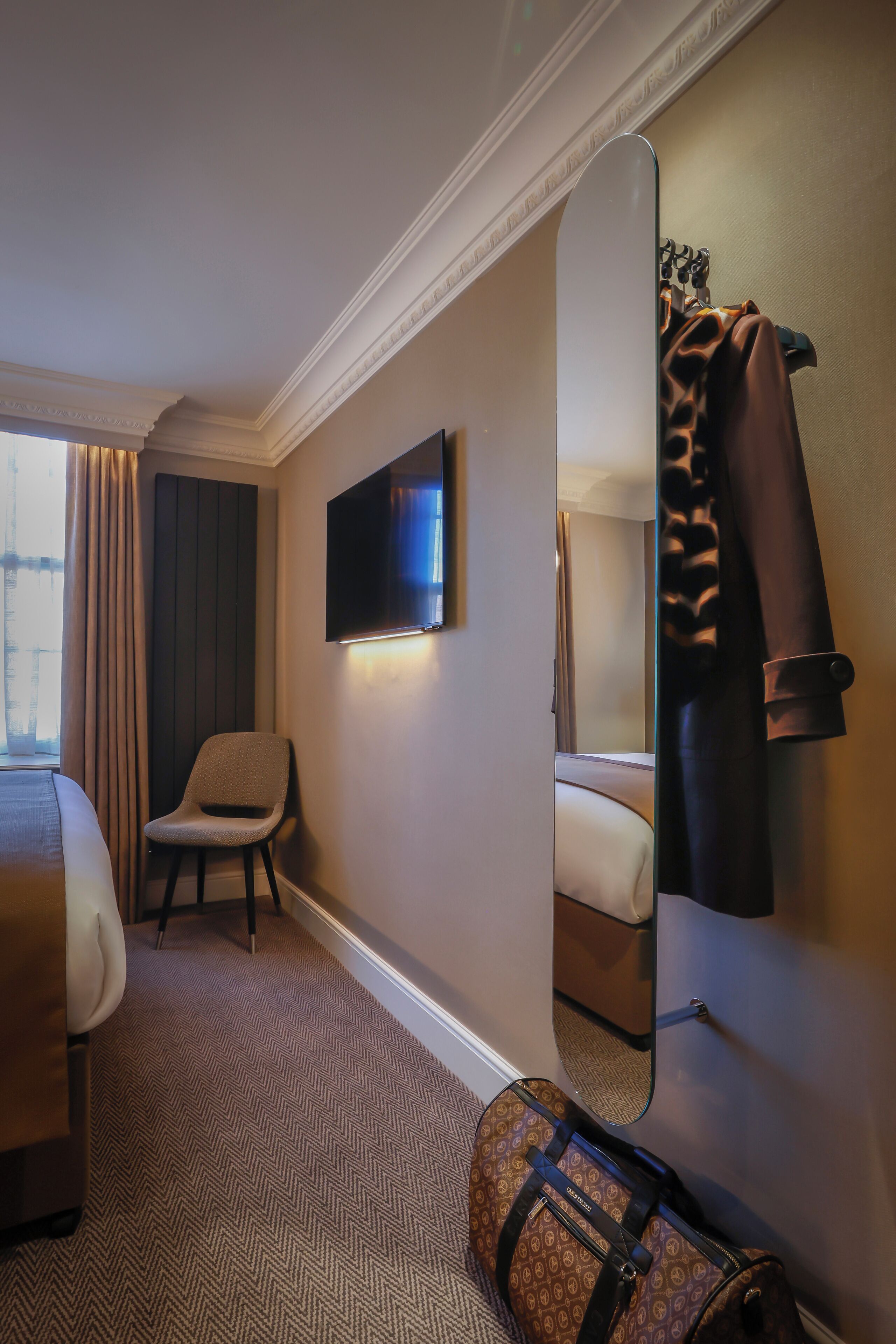 A hotel room with a bed, a chair, a television, a mirror, and a suitcase.