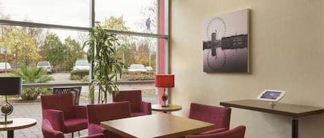 A hotel room with a large window, a table, four chairs, a lamp, and a wall-mounted tablet.