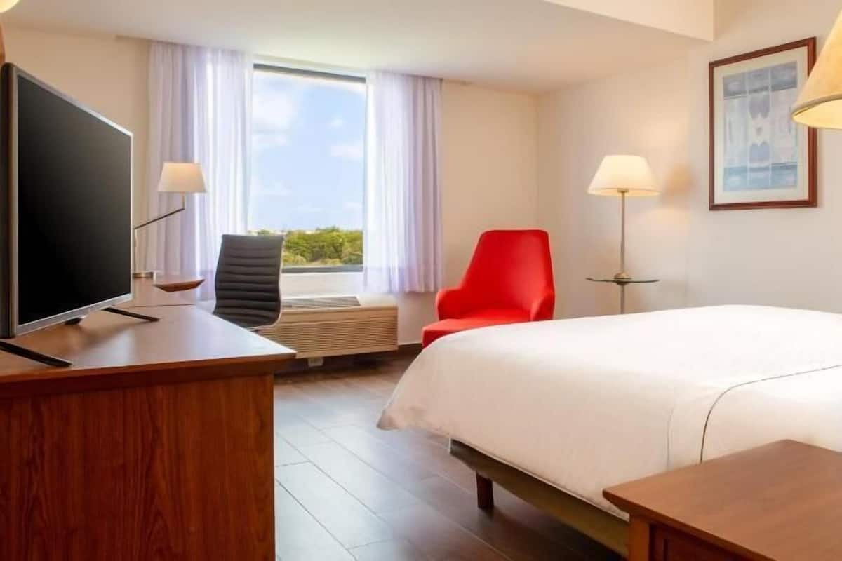 Habitació d'hotel amb llit, escriptori, cadira, televisió, làmpada i finestra amb cortines.