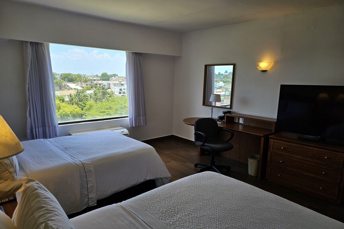 A hotel room with two beds, a desk, a chair, a TV, and a window with curtains.