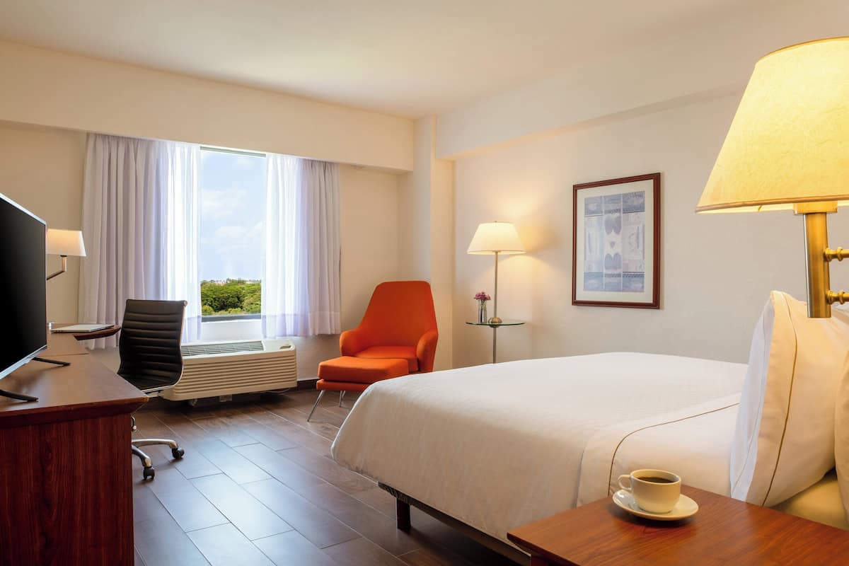 Habitació d'hotel amb un llit, una taula de treball amb un ordinador, una cadira taronja, una finestra amb cortines, una làmpada i un quadre al mur.