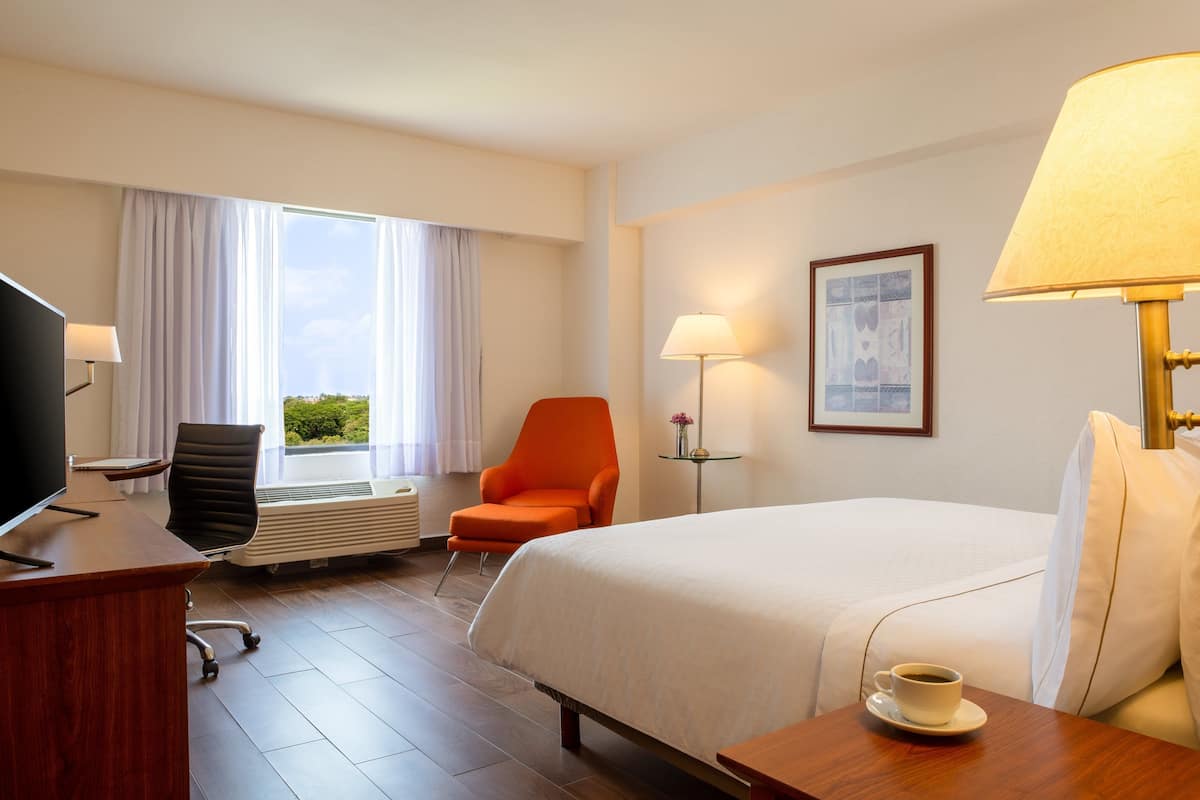 Habitació d'hotel amb llit, escriptori amb televisió, una cadira taronja, taula amb una tassa i finestra amb cortines.