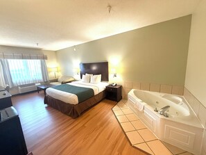 A hotel room with a large bed, a sitting area with a chair and table, a desk, a large bathtub, and a window with curtains.