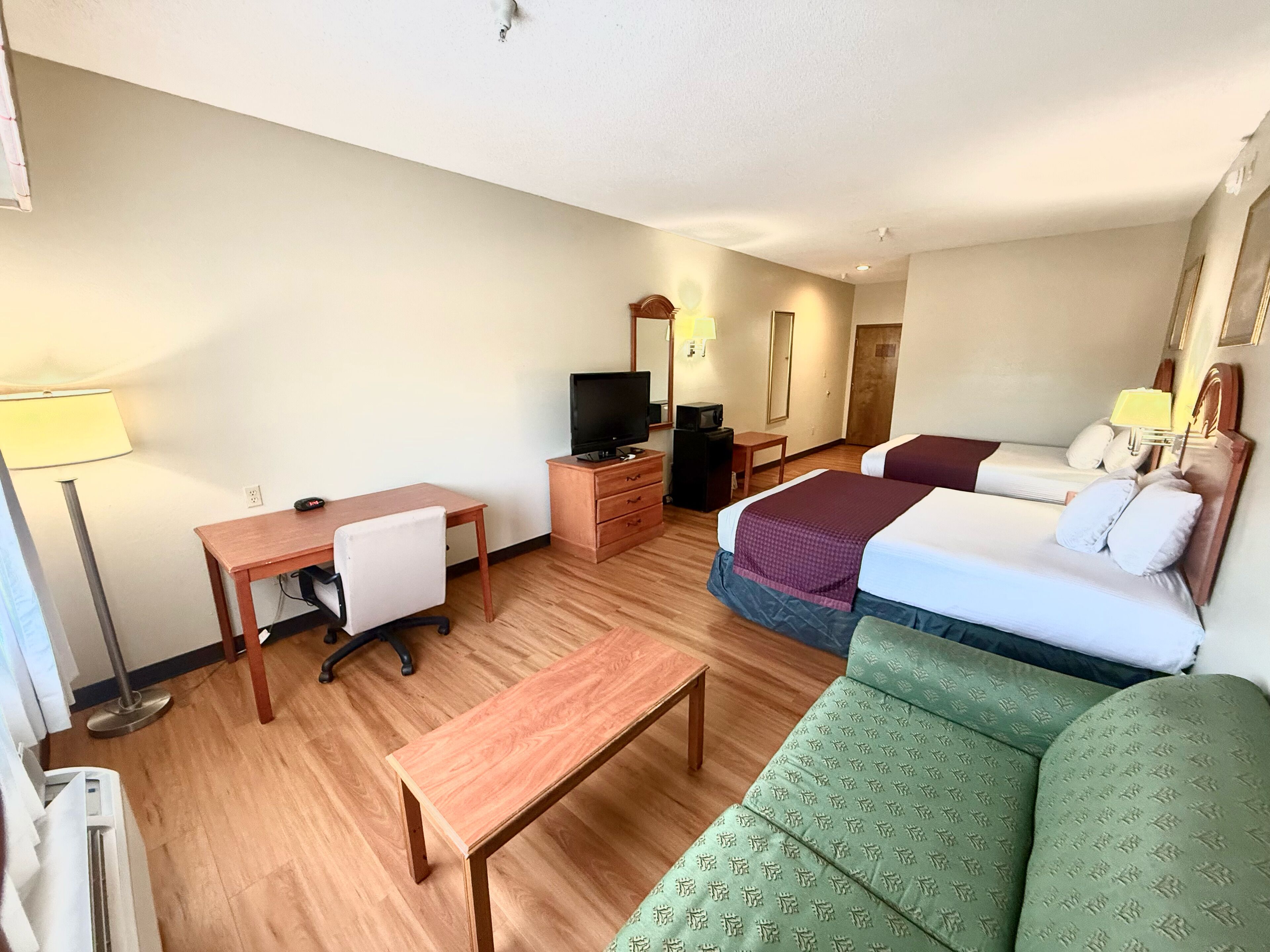 Ein Hotelzimmer mit Holztisch, einem grünen Sofa, zwei Betten mit Kissen, einem Fernseher, einem Schminktisch und einer Lampe.